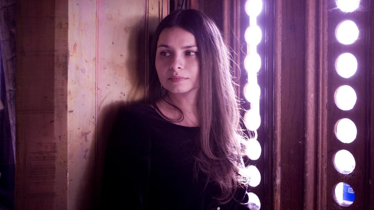 Hope Sandoval posing for a portrait, wearing a vintage-style dress and looking away from the camera.