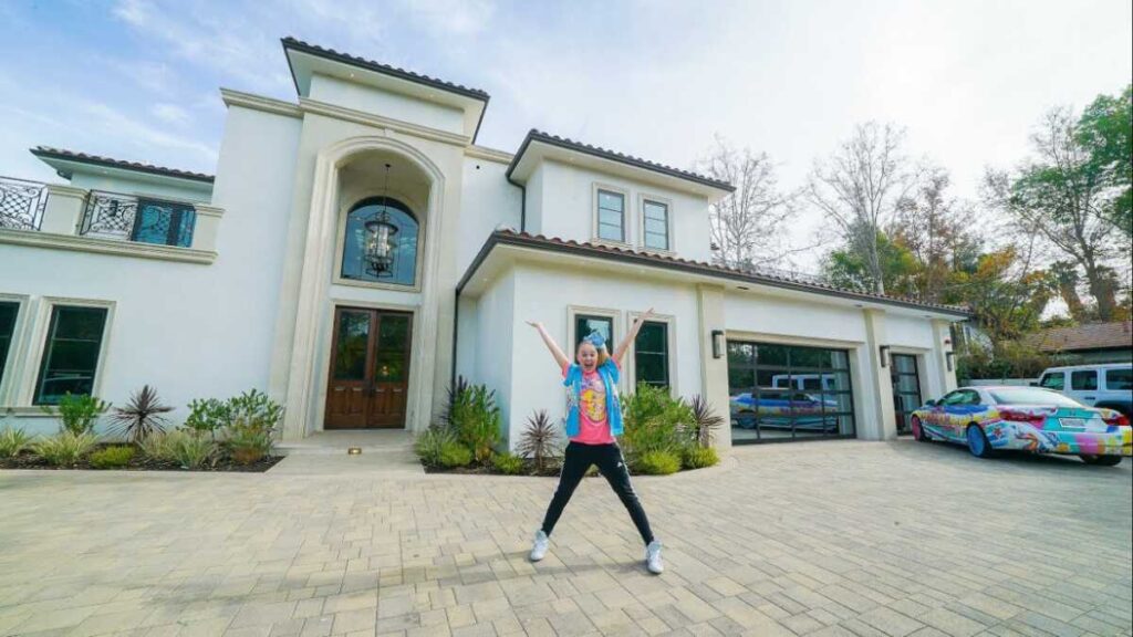 JoJo Siwa posing in front of her colorful Los Angeles mansion featuring luxury cars and vibrant decor, reflecting her bold style and multimillion-dollar lifestyle in 2025.