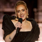 Adele performing live on stage, wearing a black gown and holding a microphone.