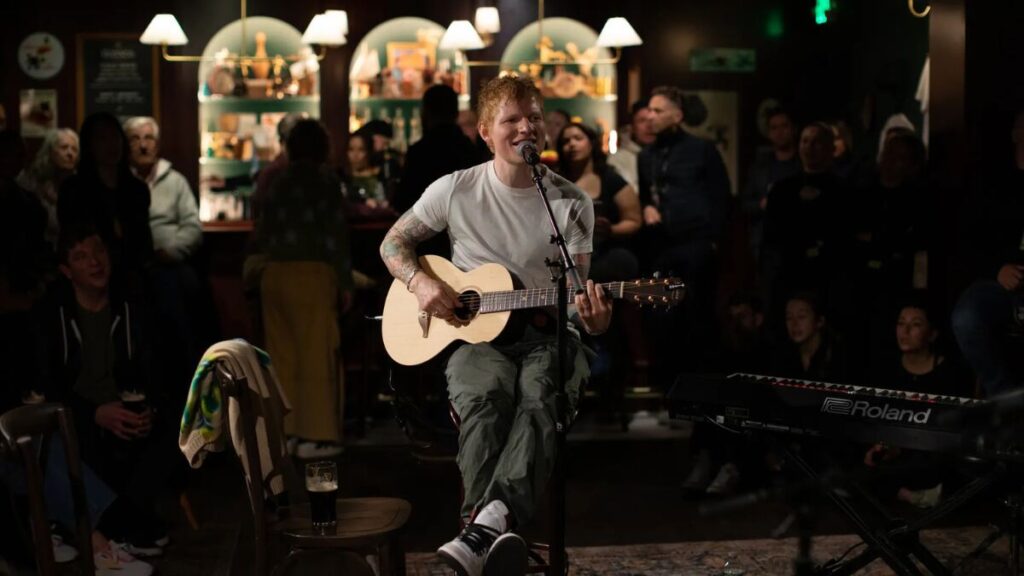 Ed Sheeran performing an intimate acoustic set in a hotel lounge, surrounded by seated audience members.