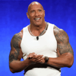 Dwayne “The Rock” Johnson standing on stage in a sleeveless shirt, showcasing his muscular build and charismatic presence.