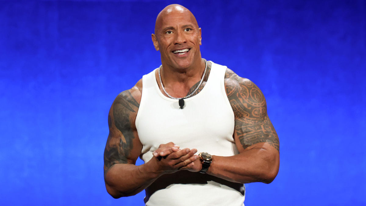 Dwayne “The Rock” Johnson standing on stage in a sleeveless shirt, showcasing his muscular build and charismatic presence.