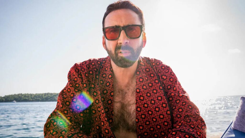 Nicolas Cage posing during a seaside photo shoot, dressed in casual coastal attire with waves and rocky shoreline in the background.