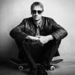 Tony Hawk posing in a black suit while seated on skateboard, studio photo shoot