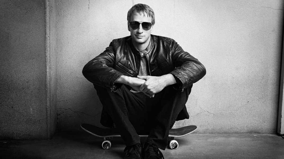 Tony Hawk posing in a black suit while seated on skateboard, studio photo shoot